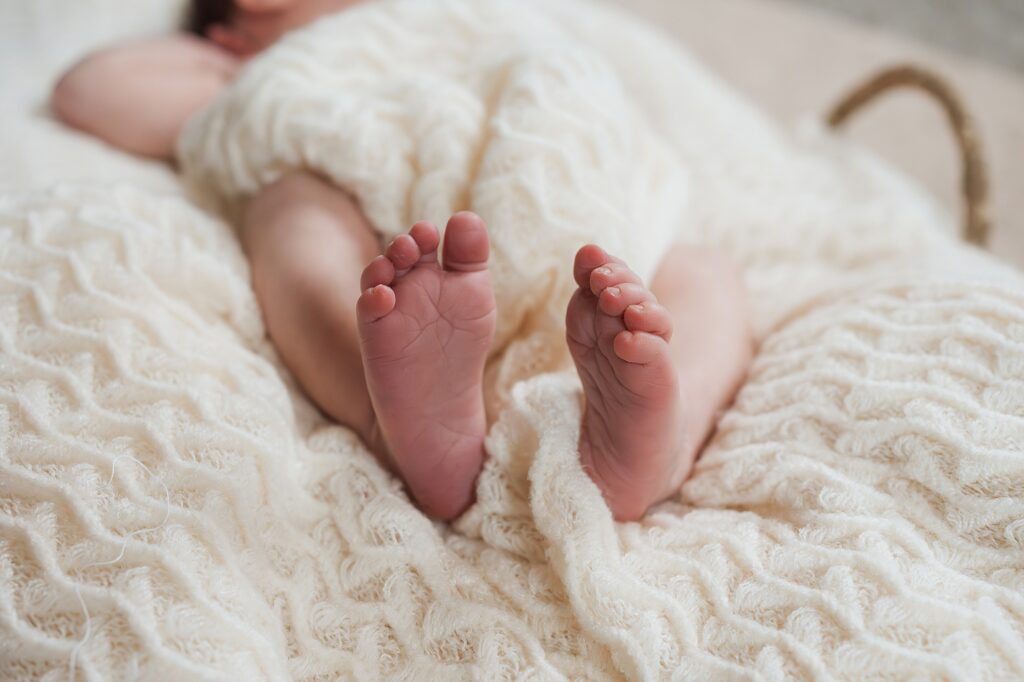 Newborn photographer captures infant's feet peeking out from a soft, cream-colored blanket.