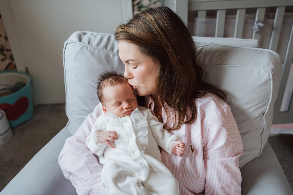 A newborn photographer captures a mother tenderly kissing her sleeping baby while sitting in a nursery.
