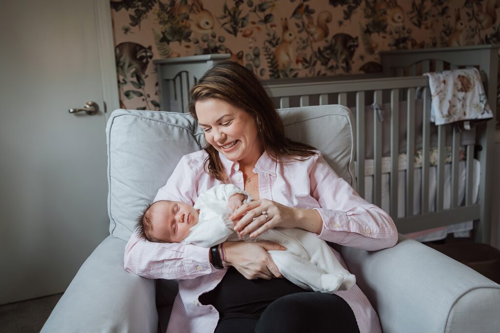 A smiling woman holding a sleeping baby in a nursery, captured by a newborn photographer.