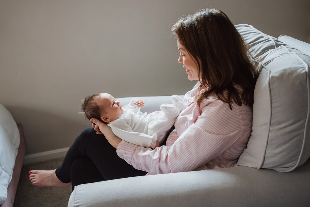 Woman sitting on a couch cradling a newborn baby, looking at the baby affectionately as a newborn photographer prepares for the shoot.