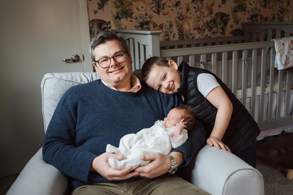 A newborn photographer sitting in a chair holding a newborn baby with a young child leaning on his shoulder.
