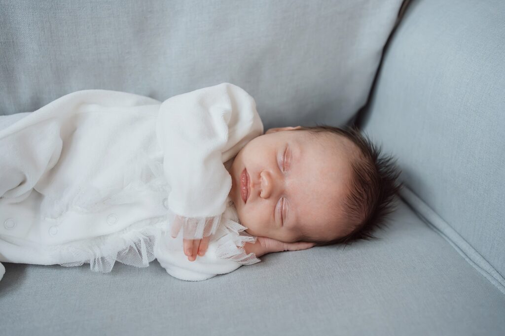 A newborn photographer captures a baby sleeping peacefully in white clothing on a grey fabric surface.