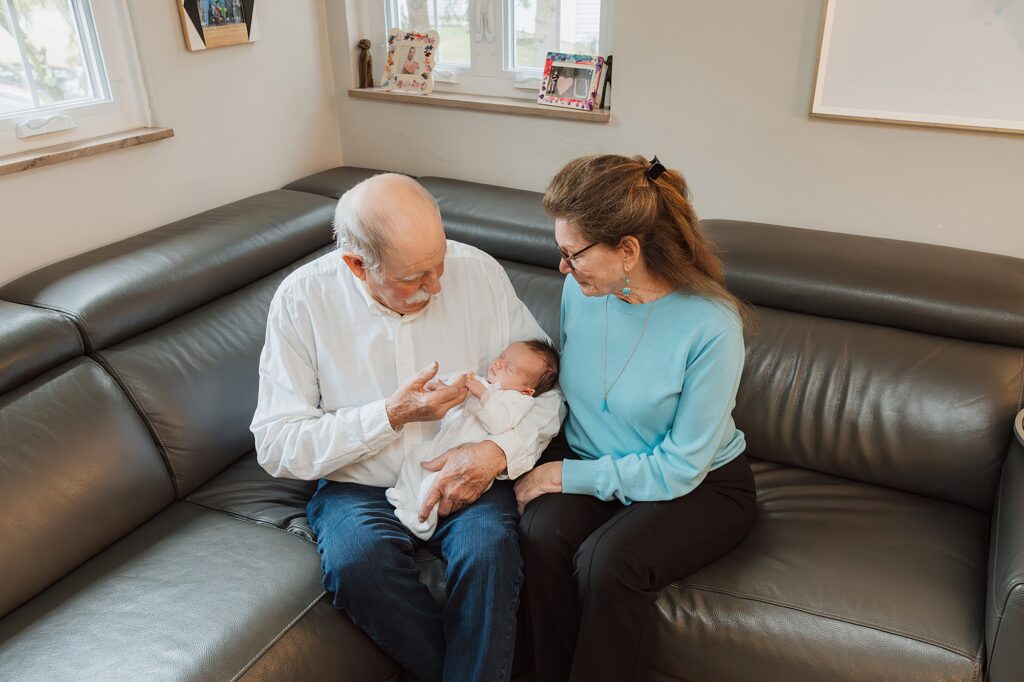 Elderly man and woman sitting on a sofa admiring a sleeping baby, as the newborn photographer sets up for the perfect shot.