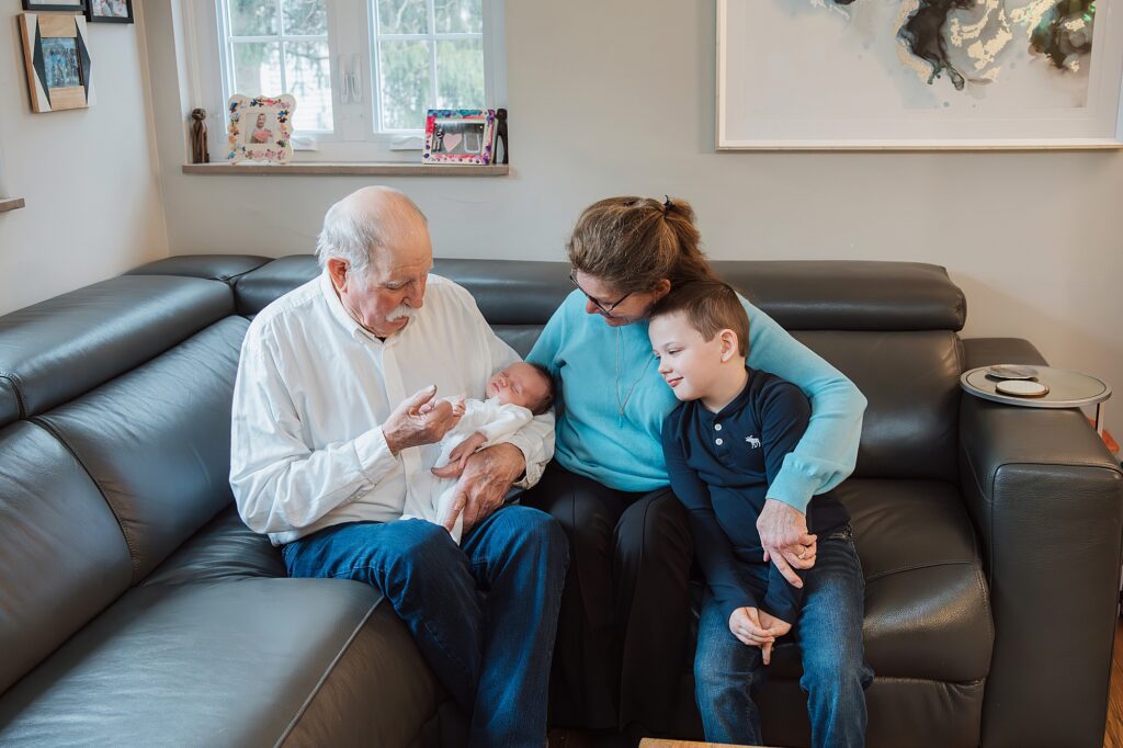 Elderly man holds a baby, woman embraces him, and young boy sits beside them on a couch, in a family home setting as the newborn photographer prepares for the shot.