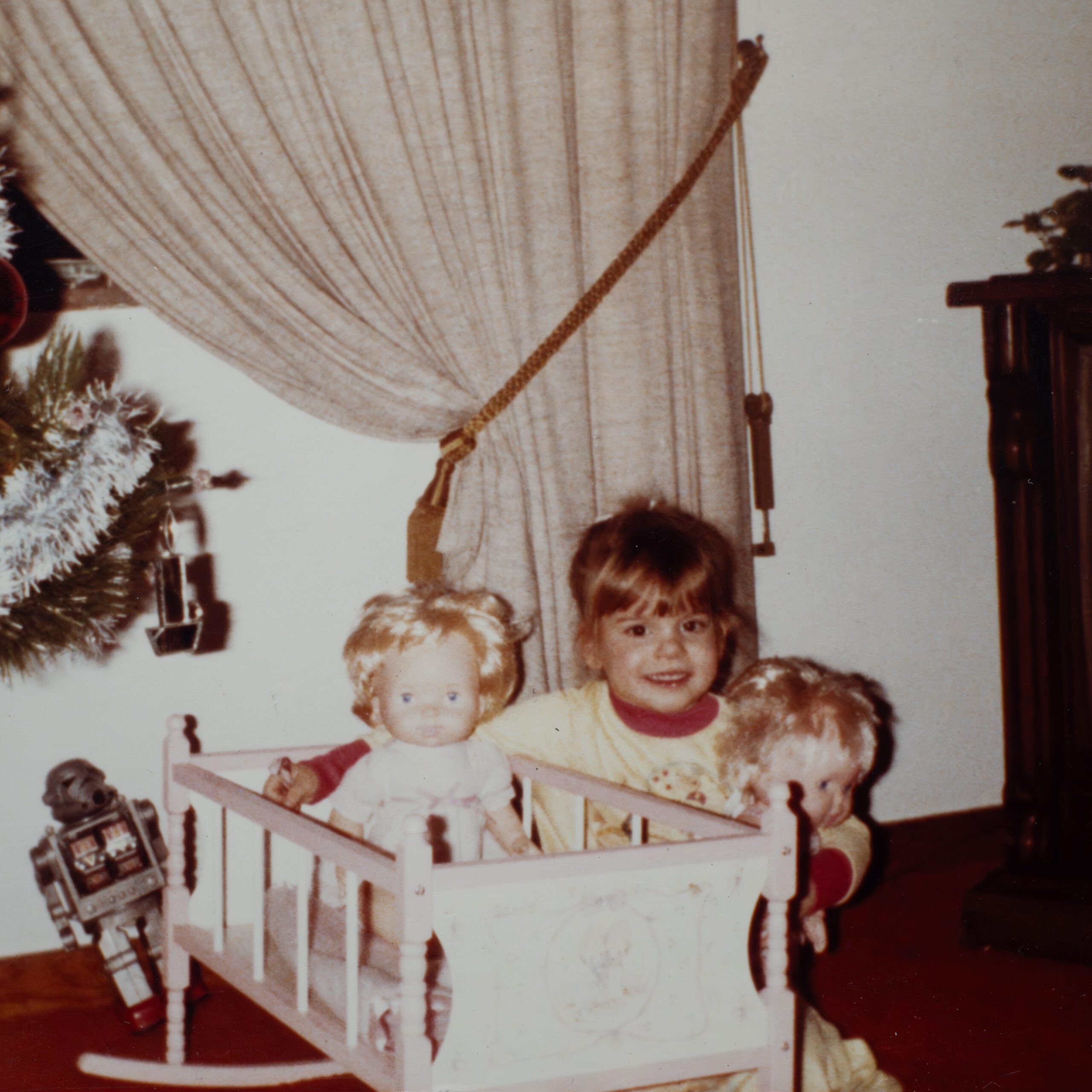 Young girl playing with dolls and a toy crib, with a decorated christmas tree in the background.