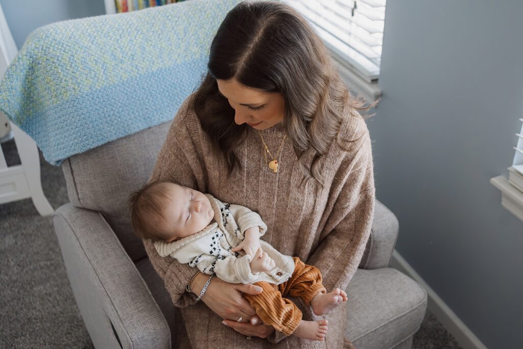 A woman sitting in a chair, gently holding and looking at a sleeping baby in a cozy room.