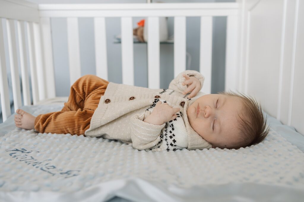 An infant sleeping peacefully in a crib while dressed in a beige cardigan and brown pants.