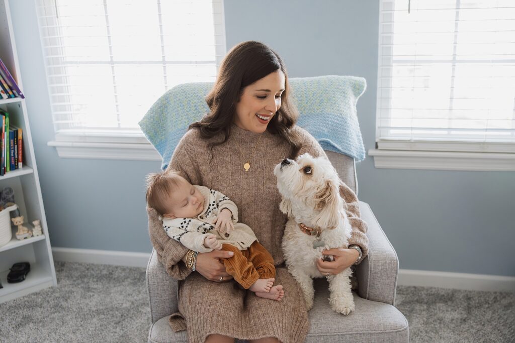 A smiling woman sitting on a chair, holding a baby and petting a small white dog.