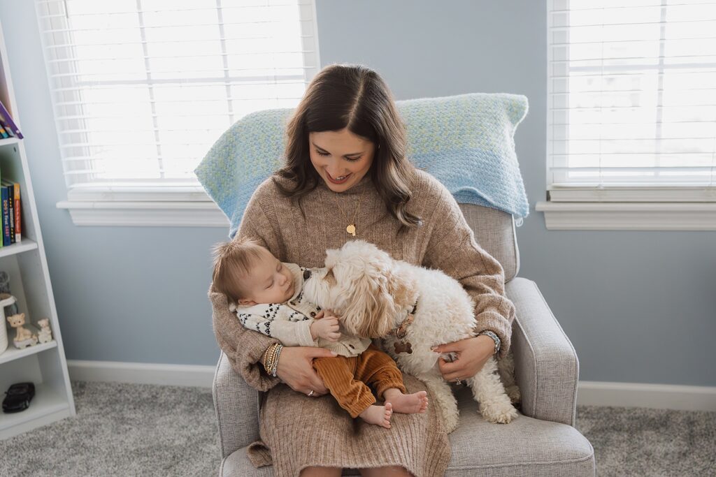 A woman sits on a chair, cradling a baby and a small dog, all enjoying a quiet moment together.