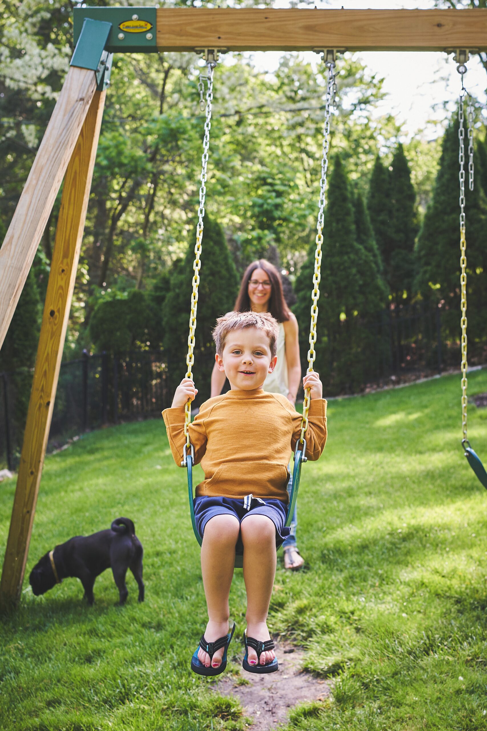 A smiling child on a swing with a woman standing behind and a small black dog in the background on a sunny day.