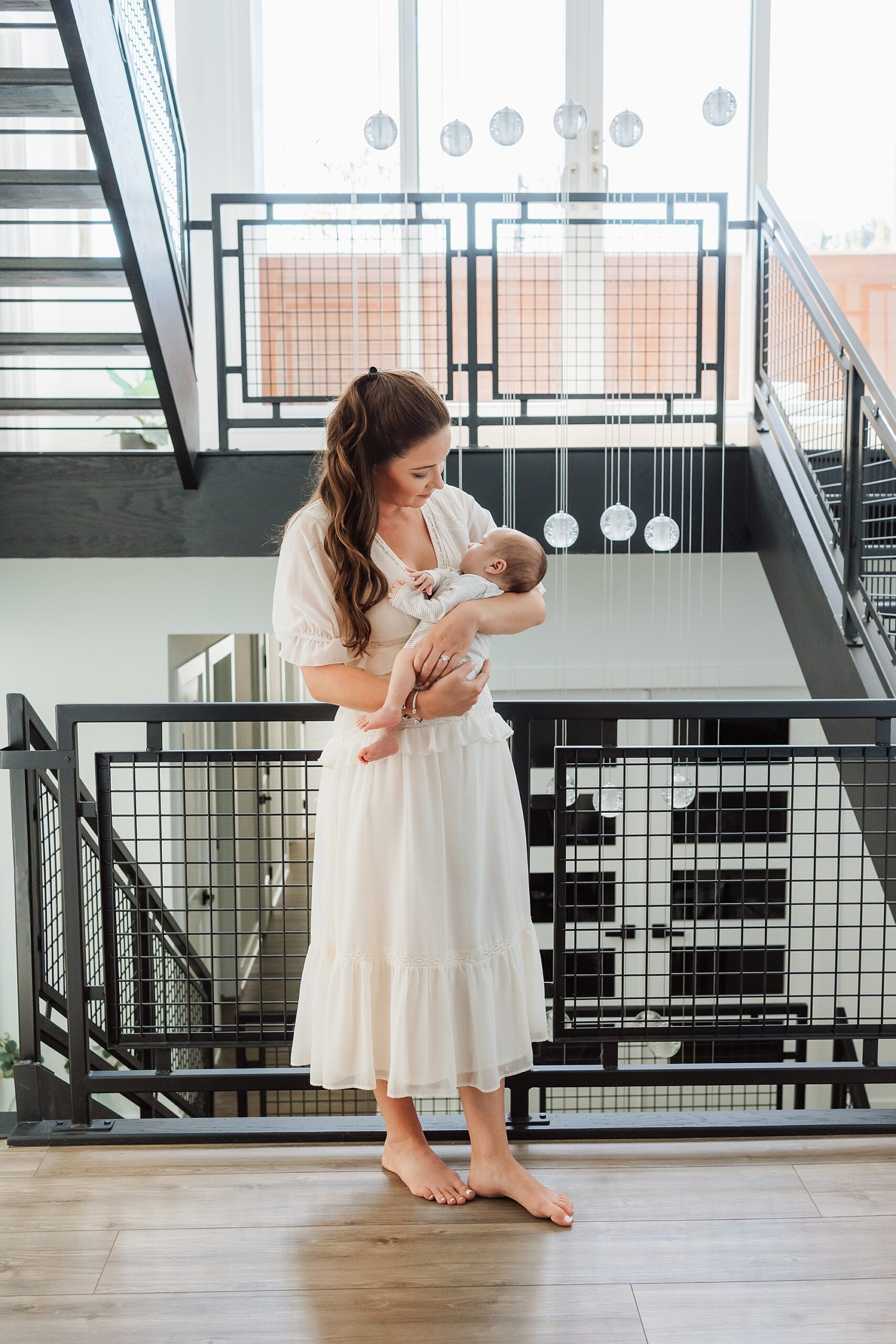 A woman in a white dress cradling a baby inside a modern home with an industrial-style staircase.