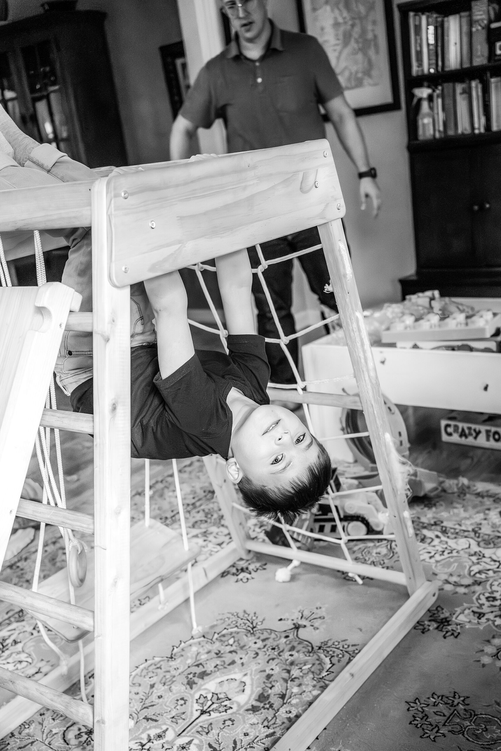 Child playfully hanging upside down from a wooden chair as an adult watches.