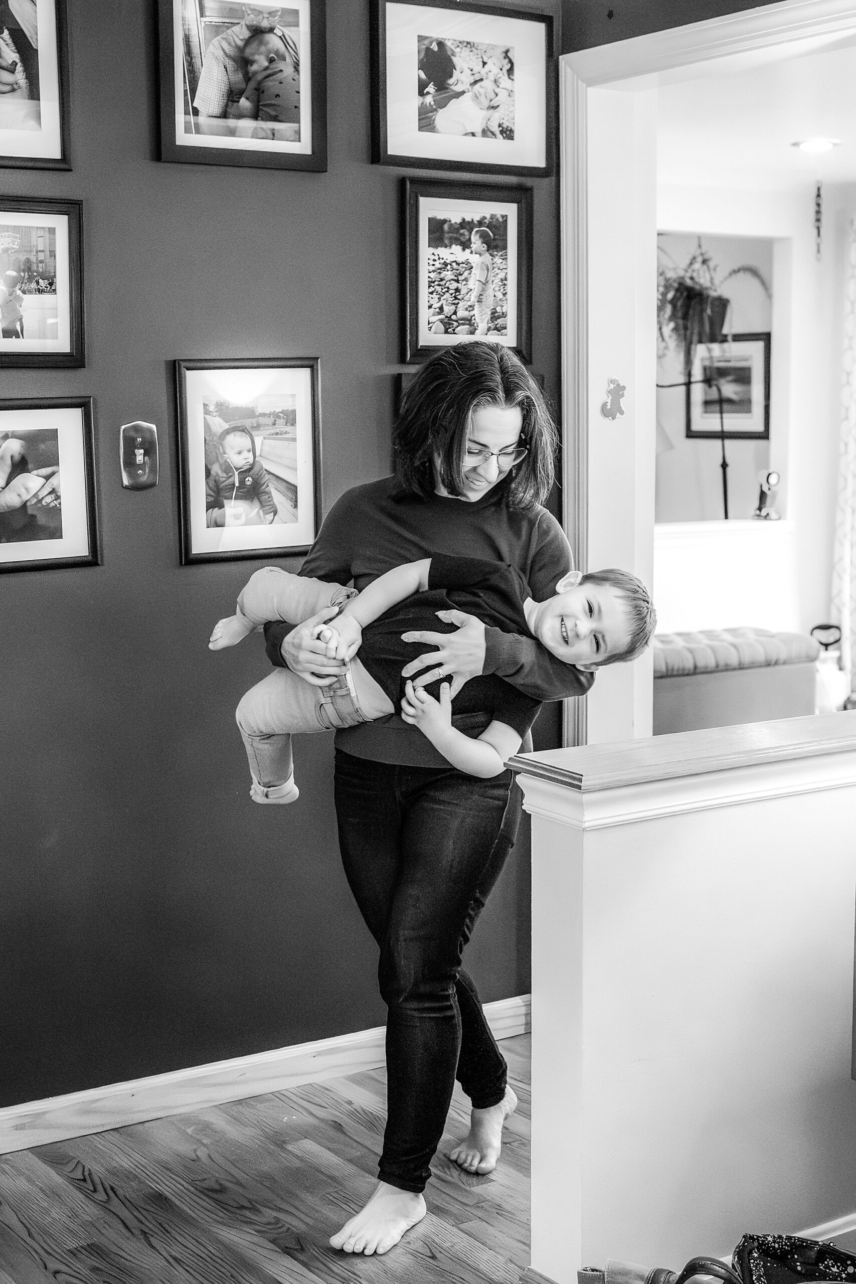 A woman carrying a playful child in a home hallway adorned with family photos.