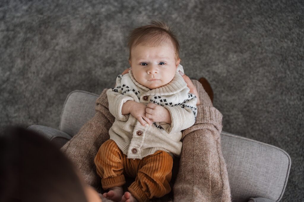 A baby with a thoughtful expression sitting on a cushioned chair, dressed in a patterned sweater and ochre pants.