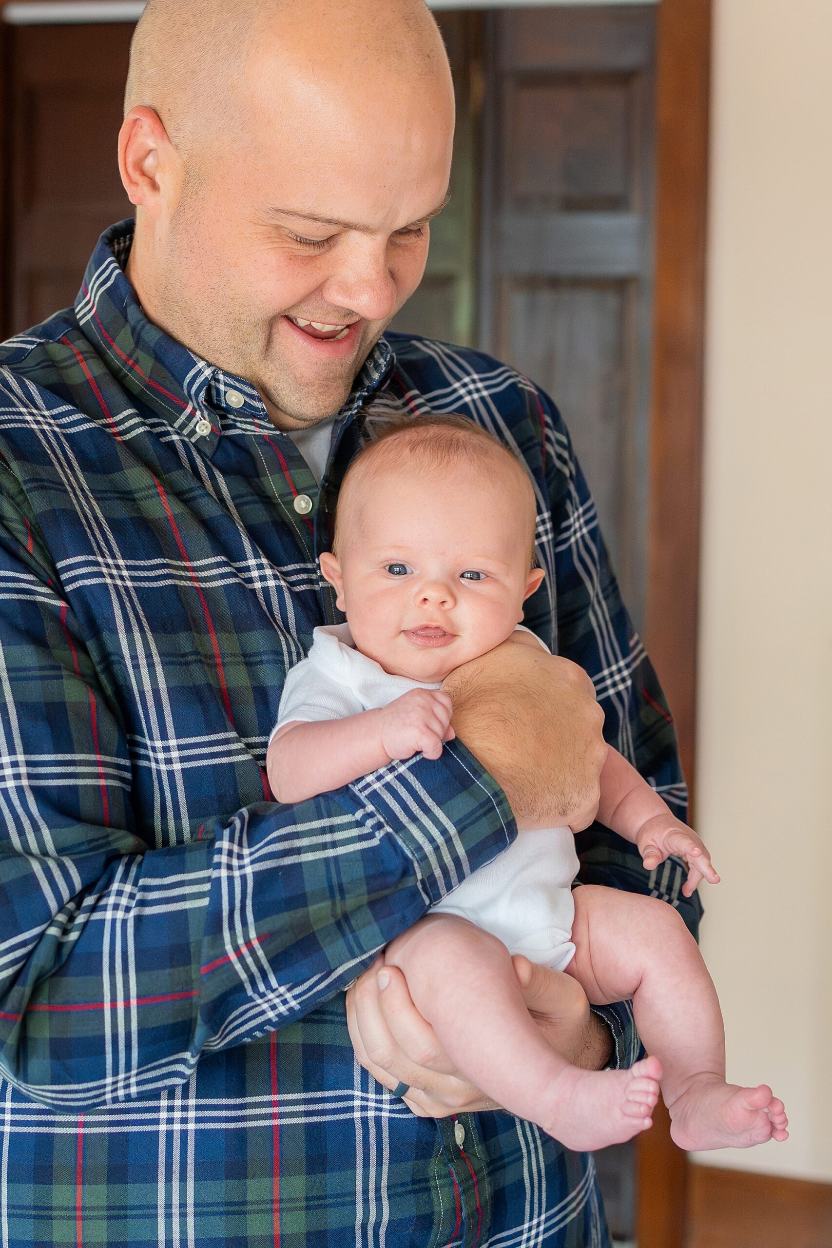 A smiling bald dad holding a baby with a slight grin indoors.