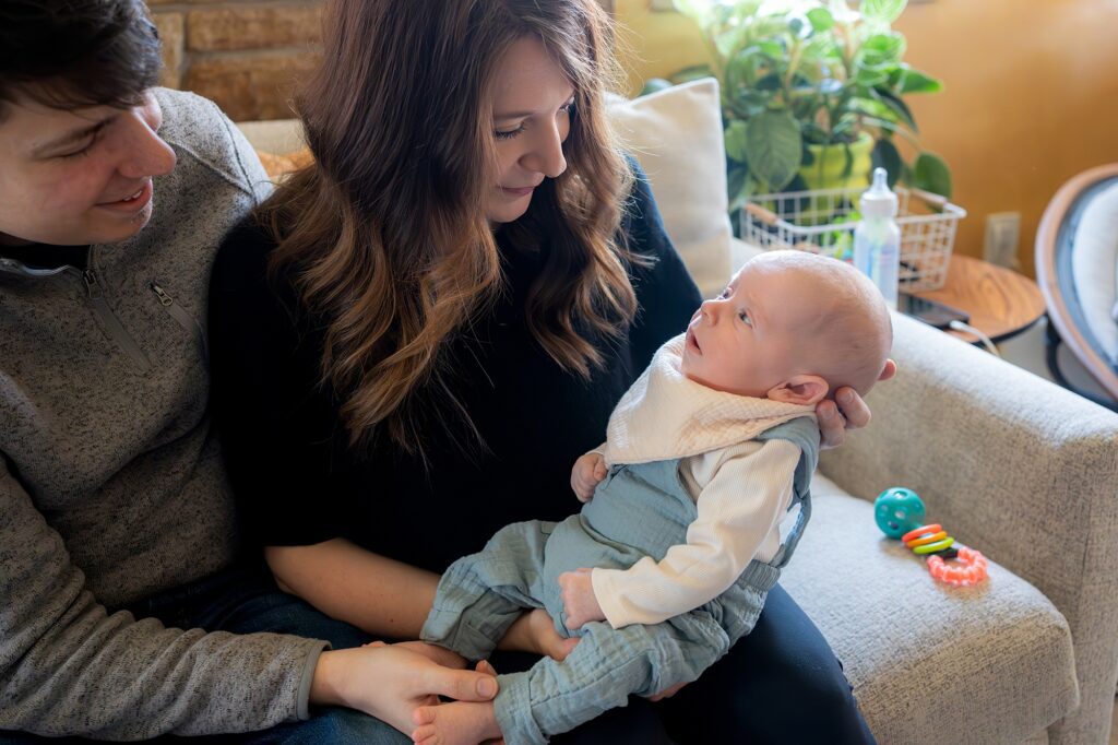 A young couple sitting on a couch, smiling lovingly at their infant son held by the mother, with baby toys nearby.