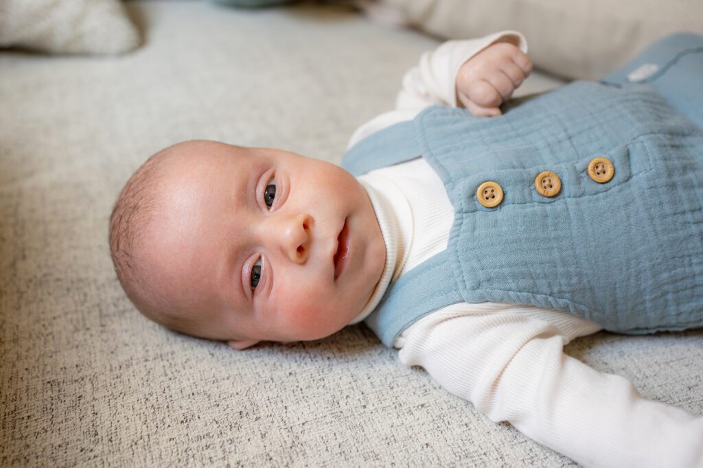 A baby wearing a blue overalls and white shirt lying on a light gray blanket, looking directly at the camera.