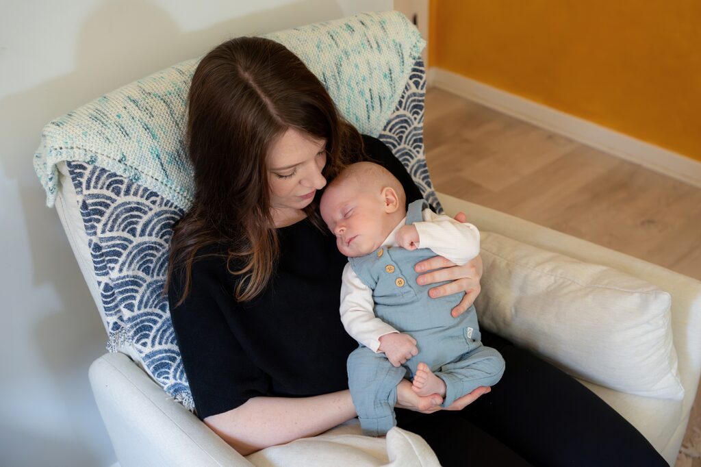 A mother tenderly holding her sleeping baby in her lap on a white armchair, with a blue and white knitted blanket draped over the back.