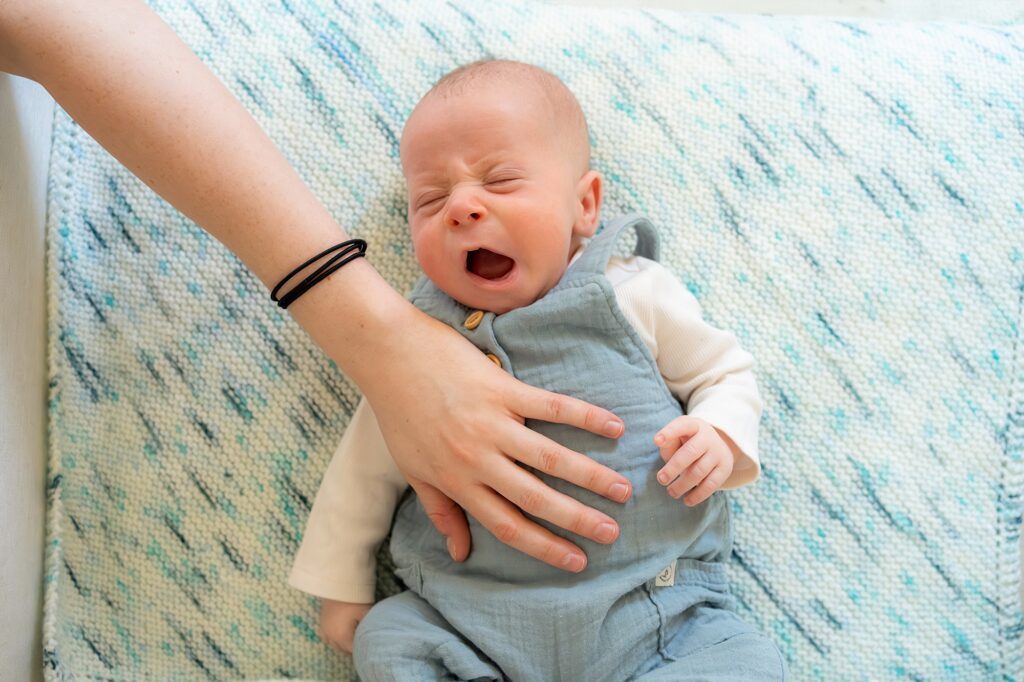 A newborn baby crying while being gently held by an adult's hand, lying on a blue and white blanket.
