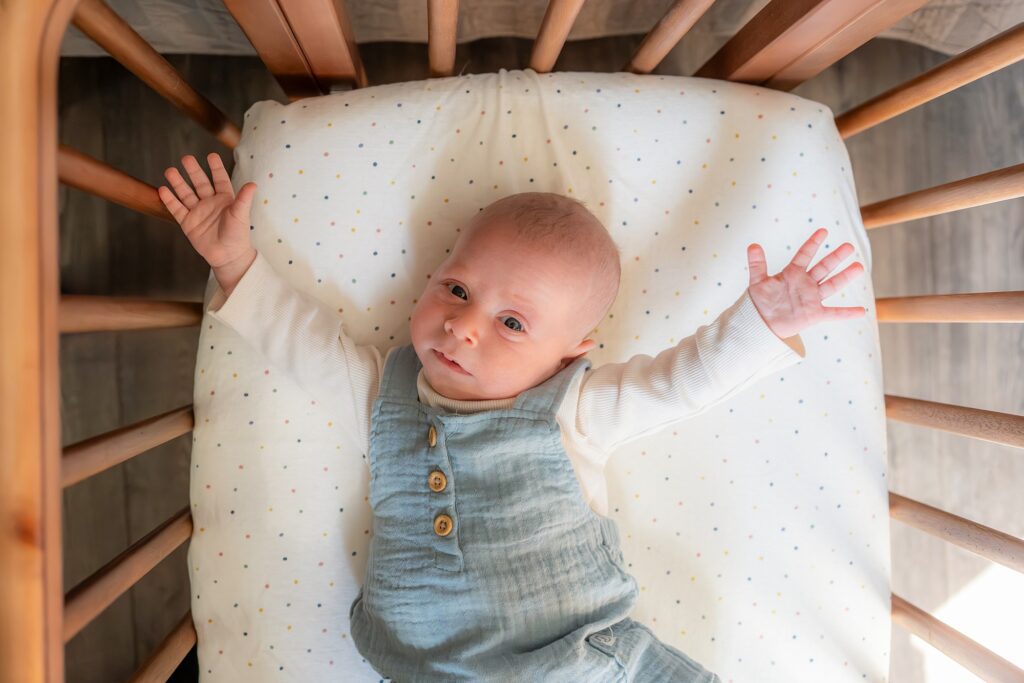 Baby wearing a green jumper lying on a polka-dot pillow in a wooden crib, looking up at the camera with arms raised.