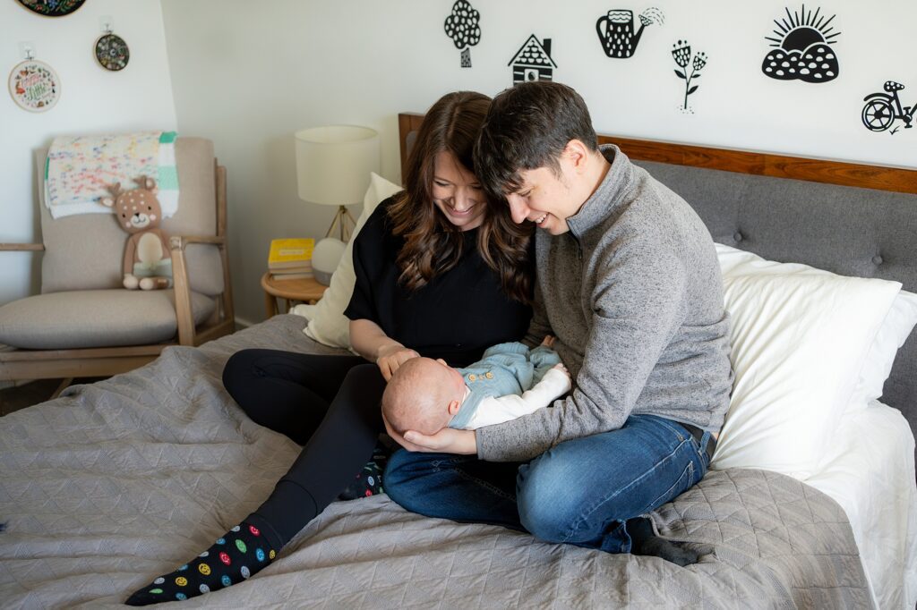 A couple sitting on a bed adoringly looking at their baby, with nursery-themed wall decorations in the background.