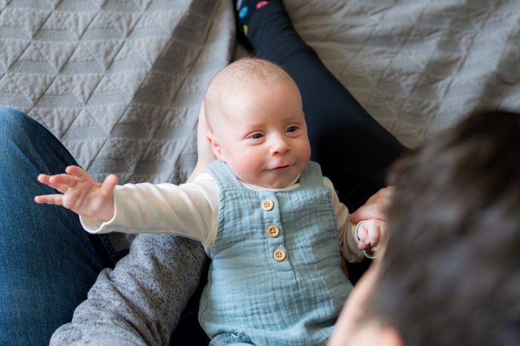 A baby wearing a striped onesie smiles and reaches out while sitting between an adult's legs on a quilted blanket.