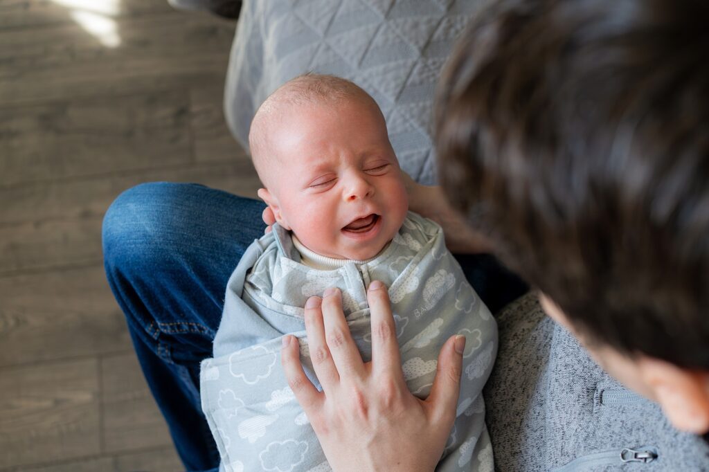 A newborn baby wrapped in a blanket cries while a person holds and comforts them in a cozy indoor setting.