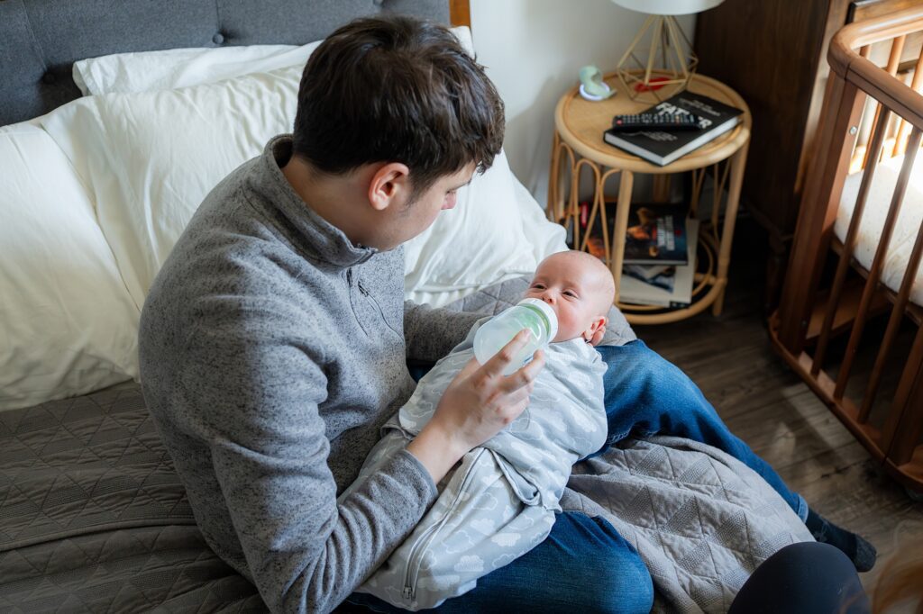 A young man feeding a baby with a bottle while sitting on a bed, with a crib nearby.