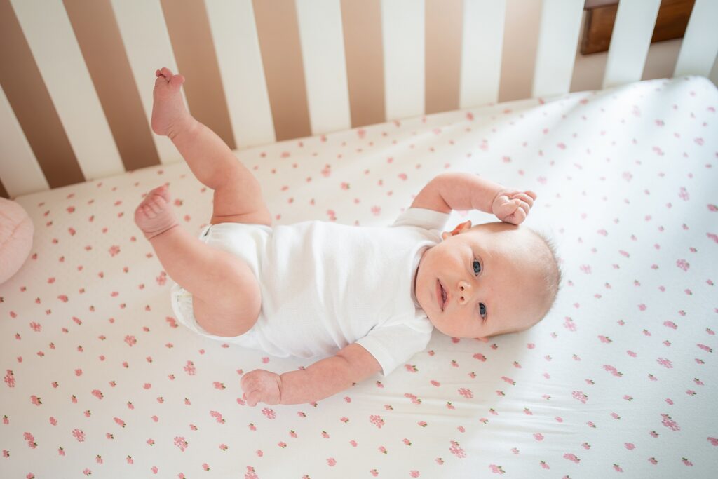 A baby lying on a crib mattress with a floral pattern, playfully kicking up legs.