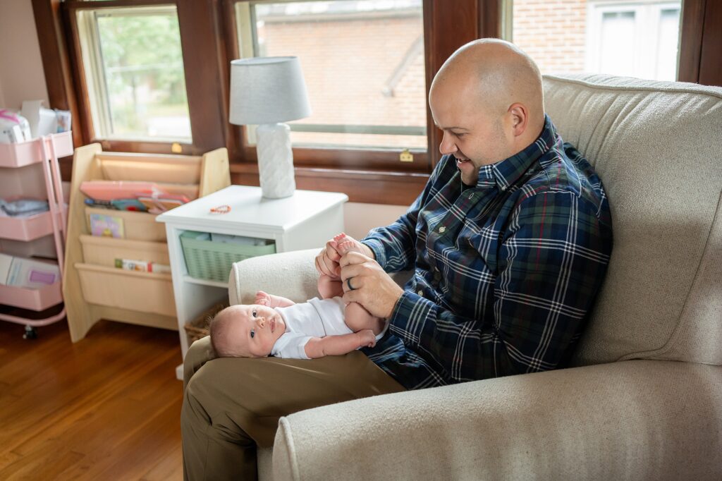 A man sitting on a sofa playfully holding a baby's feet while both of them appear to be enjoying the moment.