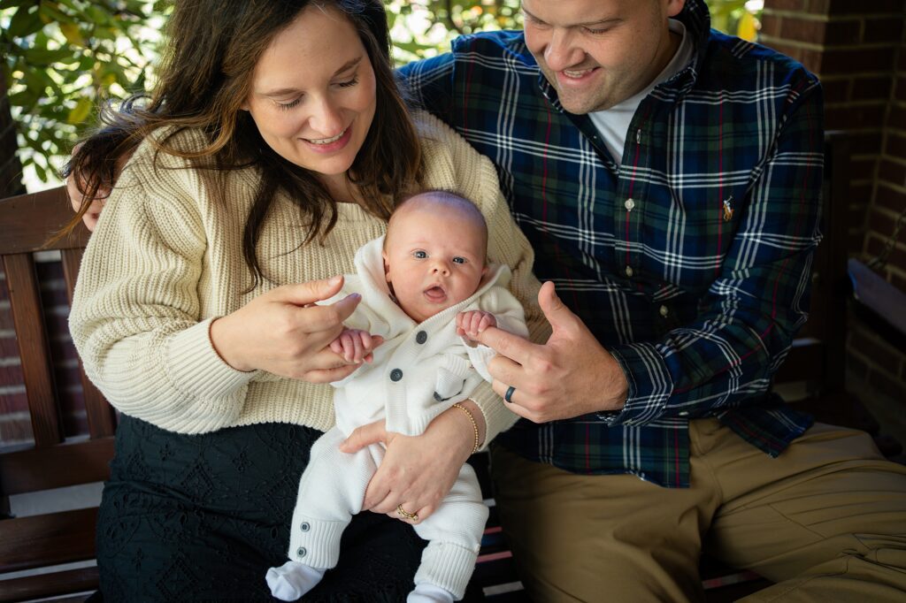 Couple sitting on a bench adoring their baby who is dressed in a white outfit.