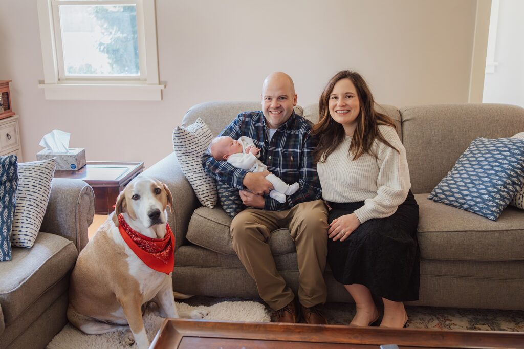 A smiling family with a newborn baby and a dog sitting on a living room couch.