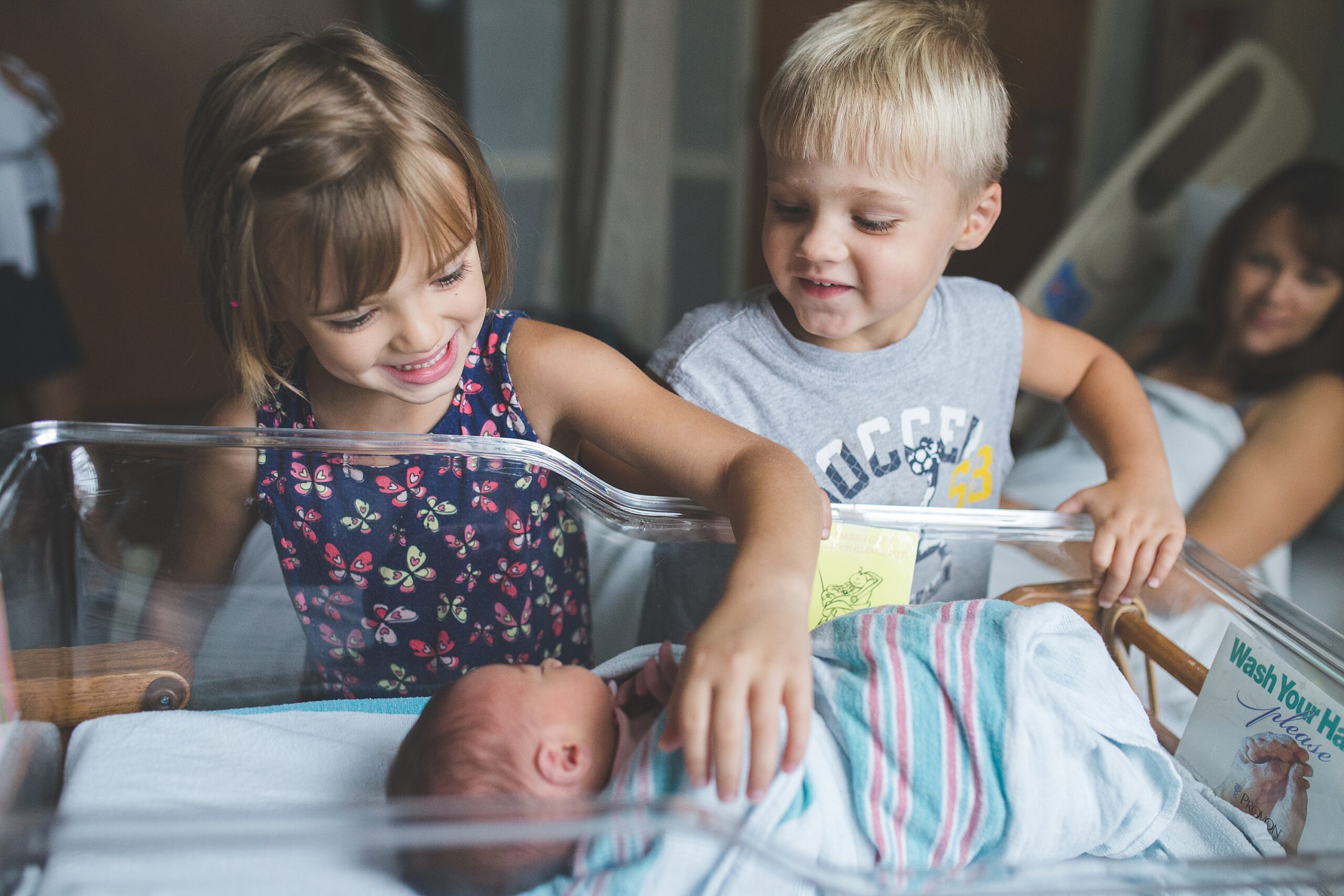 Two young children gleefully look at a newborn baby in a hospital bassinet while an adult rests in the background.