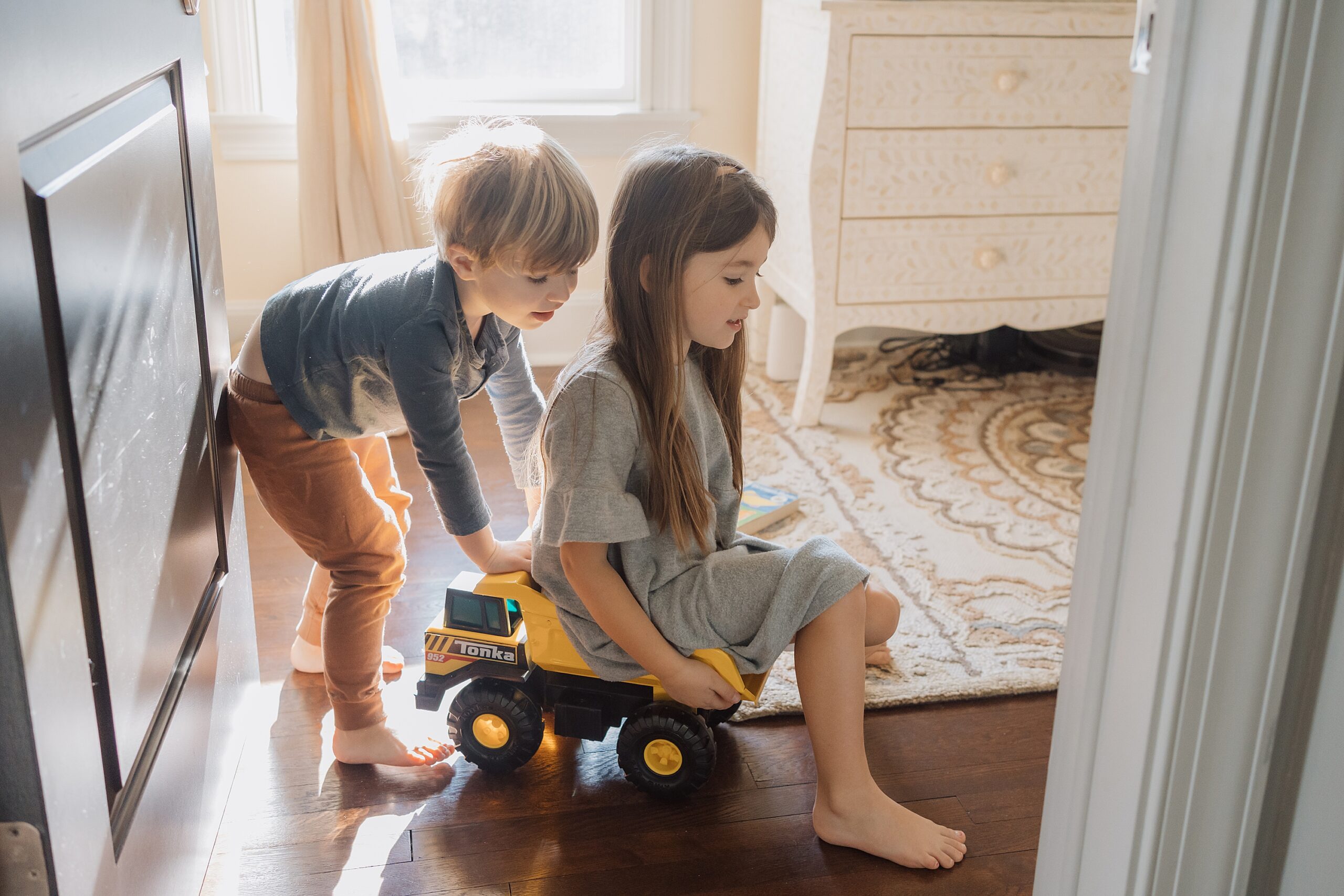 Two children playing indoors with a toy truck by a sunny window.