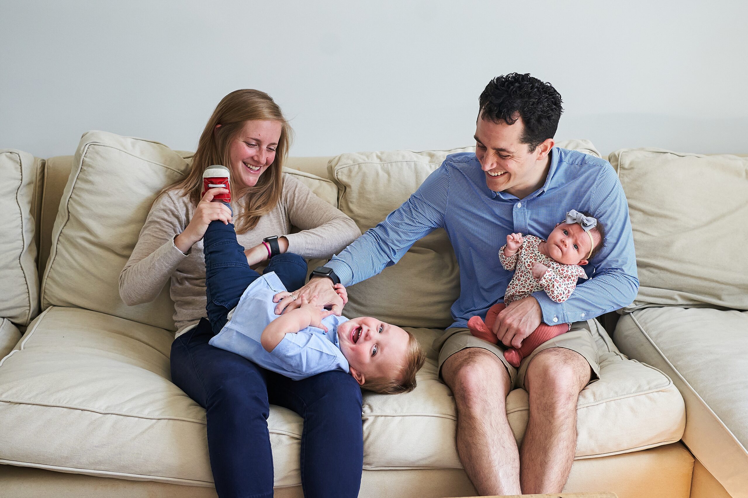 A family enjoying time together on a sofa; a mother holding a baby, a father playing with a toddler, everyone smiling.