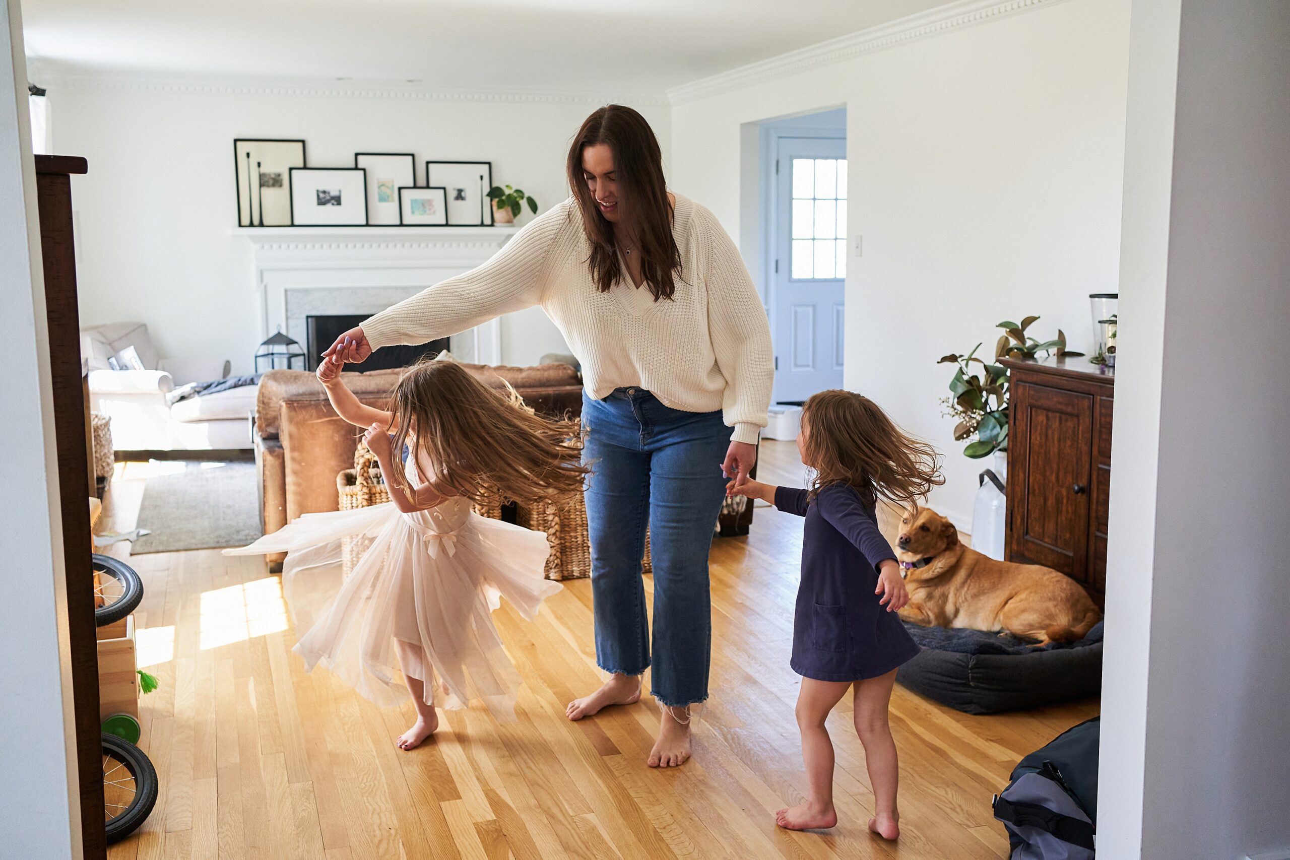 A woman plays with two young girls in a home living room while a dog watches.