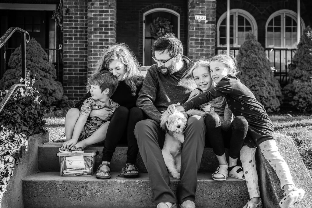 A black and white photo of a family of five and their dog sitting on the front steps of their home.