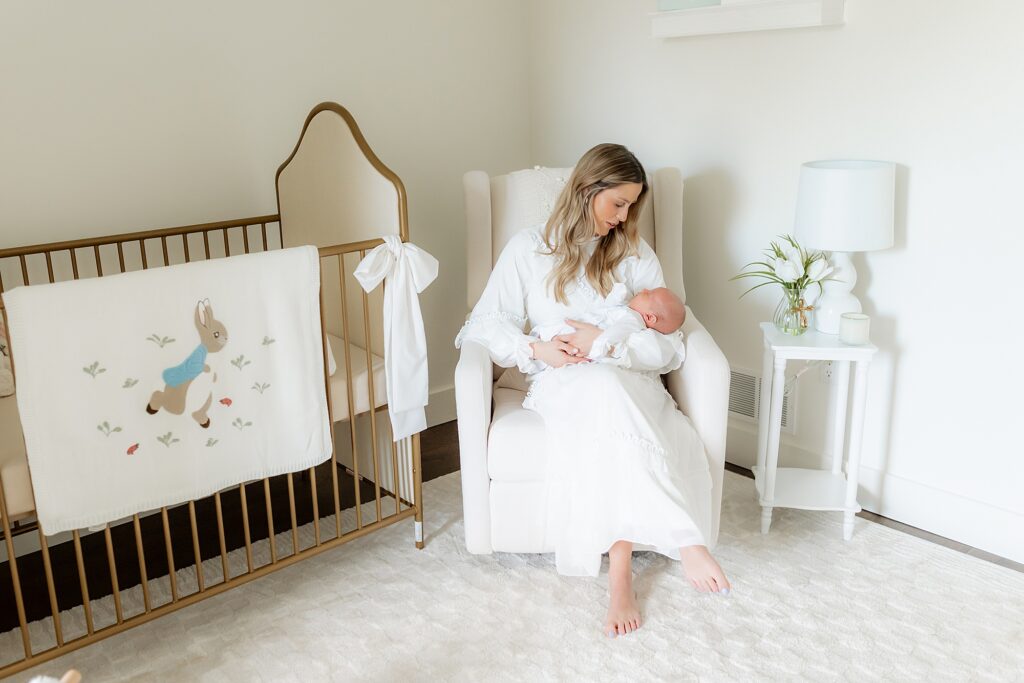 A woman in a white dress sitting on a chair, holding a newborn baby in a nursery with a crib and white decor.