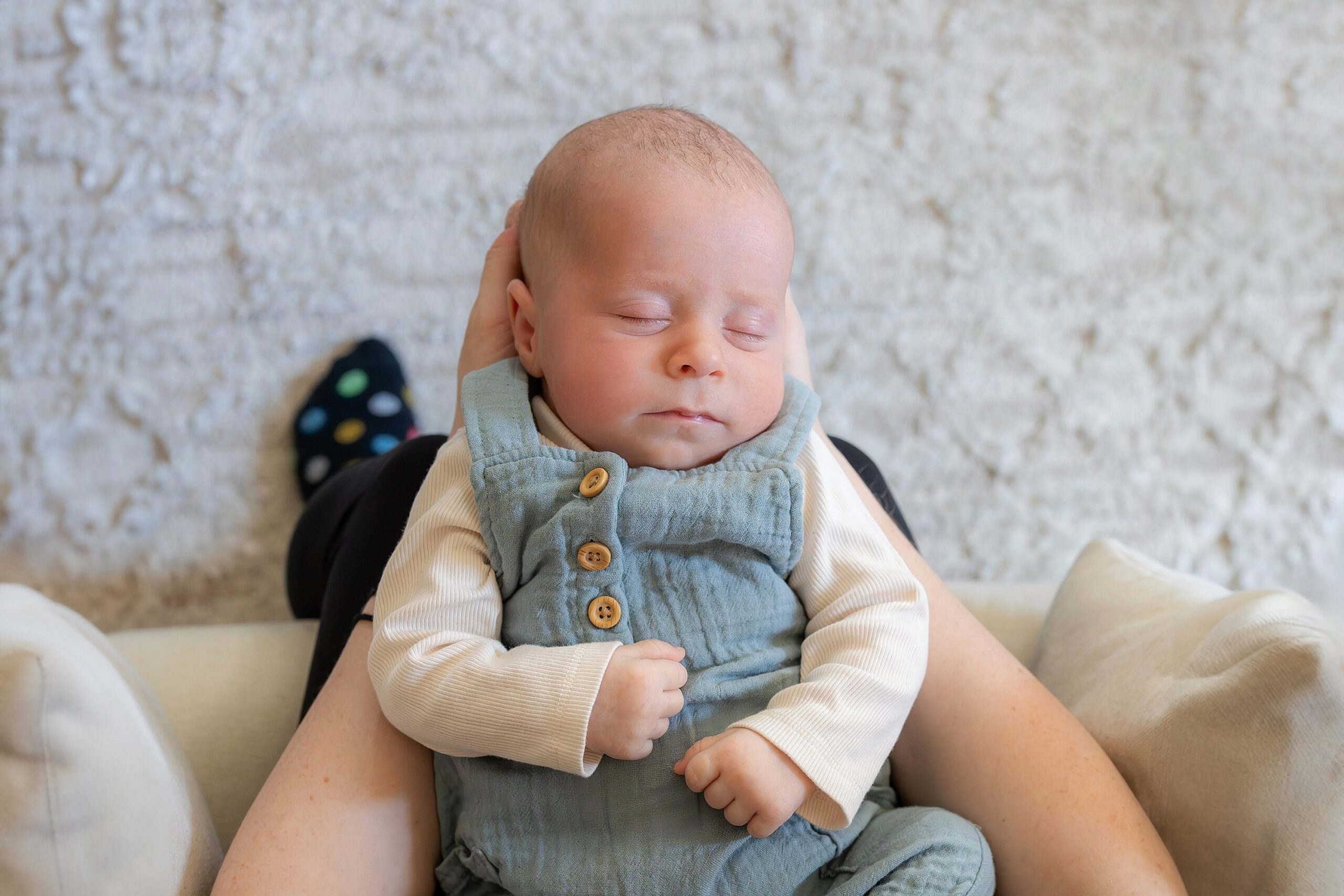 A sleeping infant in a blue onesie resting in an adult's lap against a cushioned background.