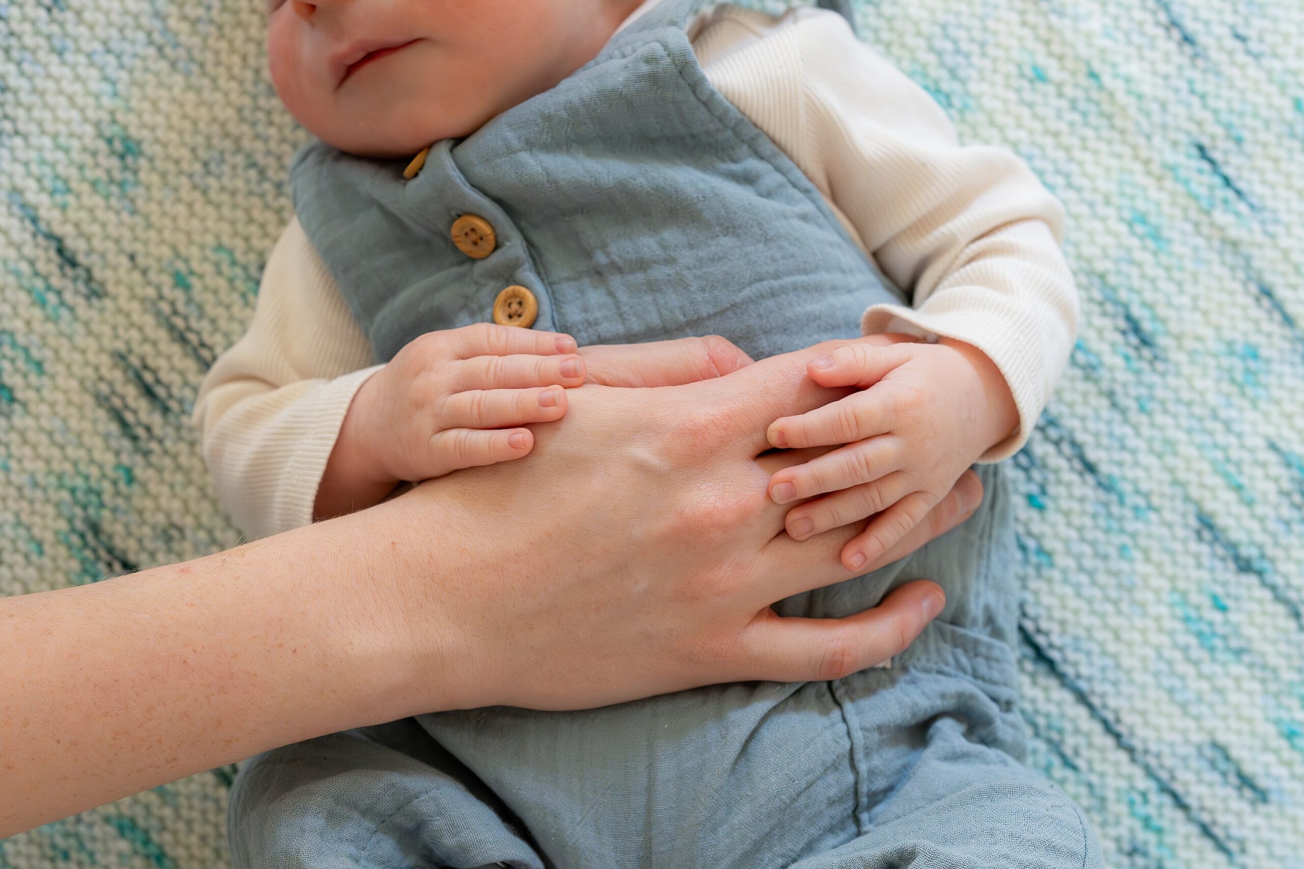 An adult's hands gently cradle a sleeping baby dressed in a blue outfit.