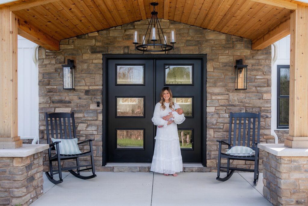 A woman in a white dress holding a baby stands at a home's entrance with a stone façade and dark double doors, flanked by two rocking chairs.