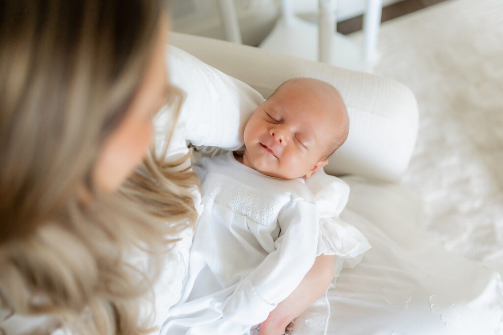 Newborn baby in a white dress sleeping on a cushion, viewed over a woman’s shoulder.