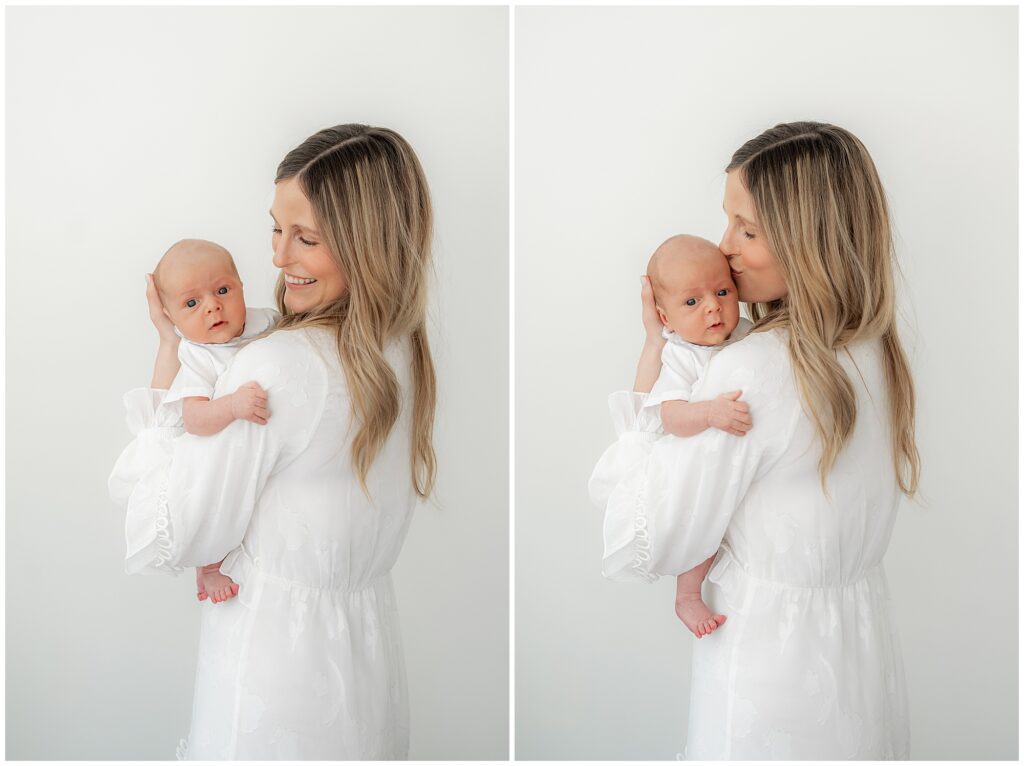 A mother in a white dress holding her newborn baby, smiling and kissing the baby's forehead in a bright room.