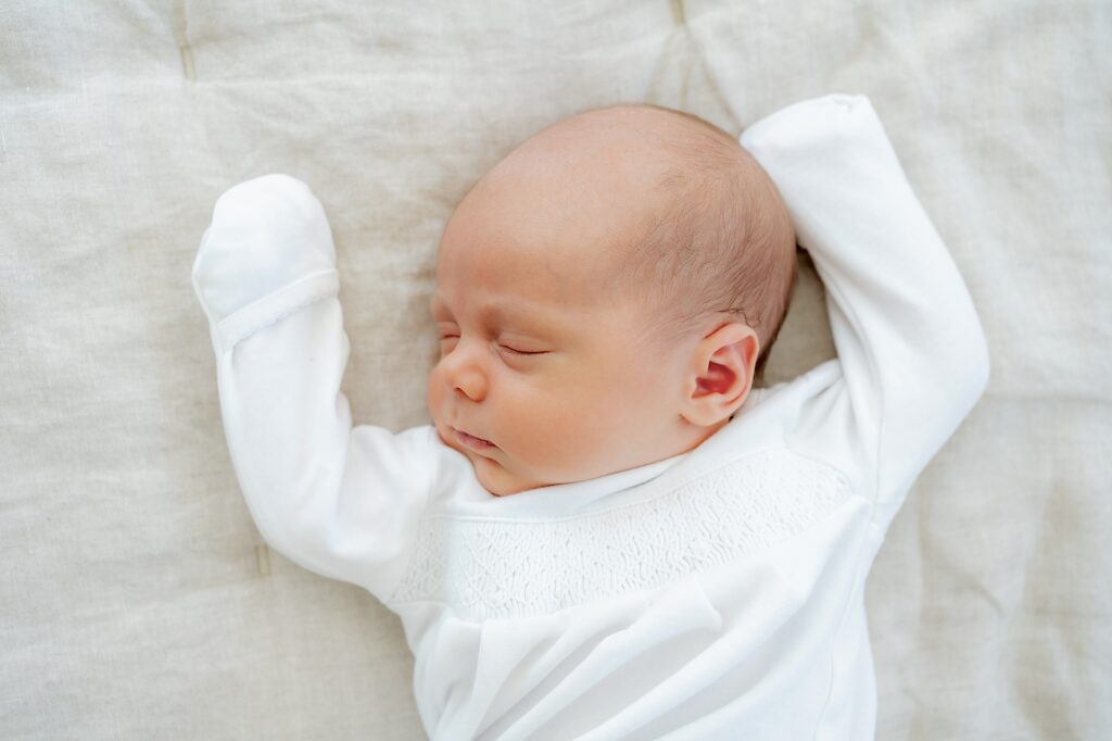 A newborn baby sleeps on a soft white blanket, dressed in a white outfit with mittens, arms raised above its head.