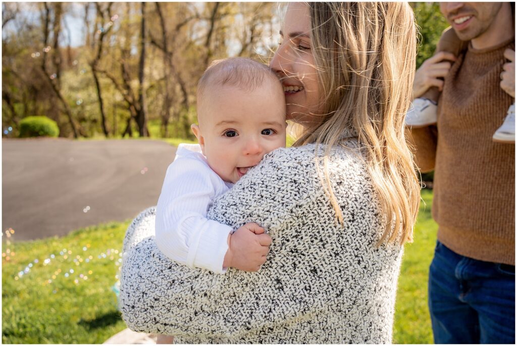 A woman holding a baby outdoors, both smiling, with a man partially visible in the background. bright, sunny day with greenery around.