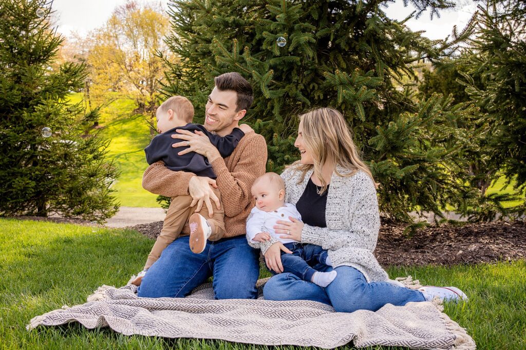 A family of four with two young children sitting on a blanket in a park, smiling and enjoying a sunny day.