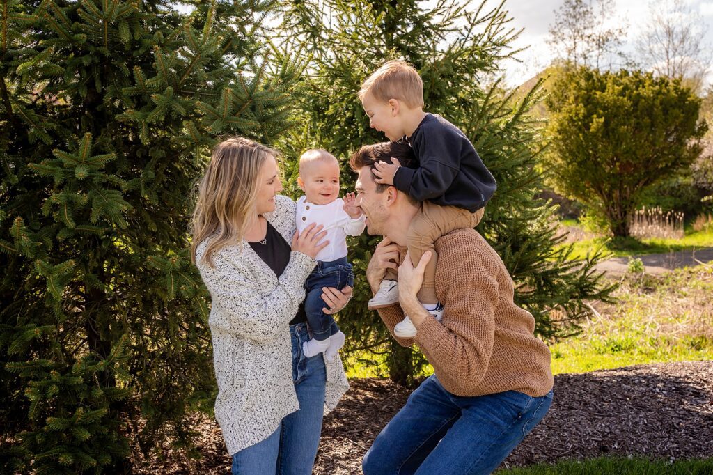 A family of four enjoying a sunny day outdoors; two parents playing with their young son and baby, surrounded by lush greenery.