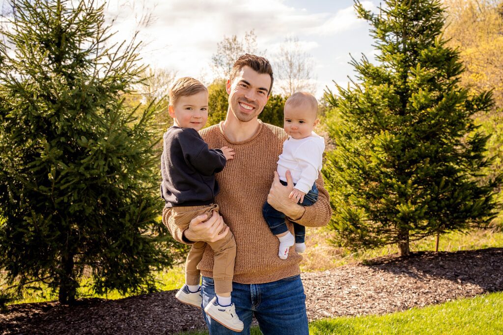 A smiling man holds a toddler on his left and a baby on his right in a sunny garden with evergreen trees in the background.