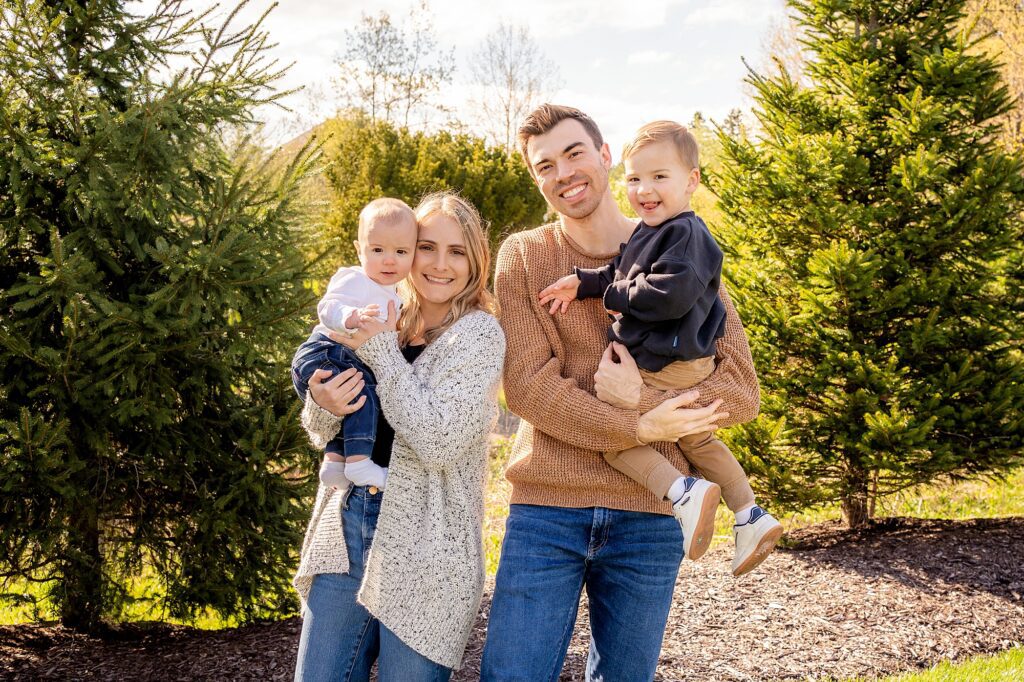 A family of four, with a couple and two young children, smiles outdoors surrounded by green trees, with the parents holding the children.