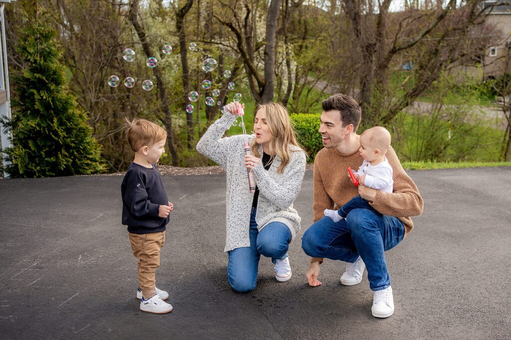 A family of four enjoying outdoors with a mother blowing bubbles towards her toddler son while the father, holding a baby, watches smiling.